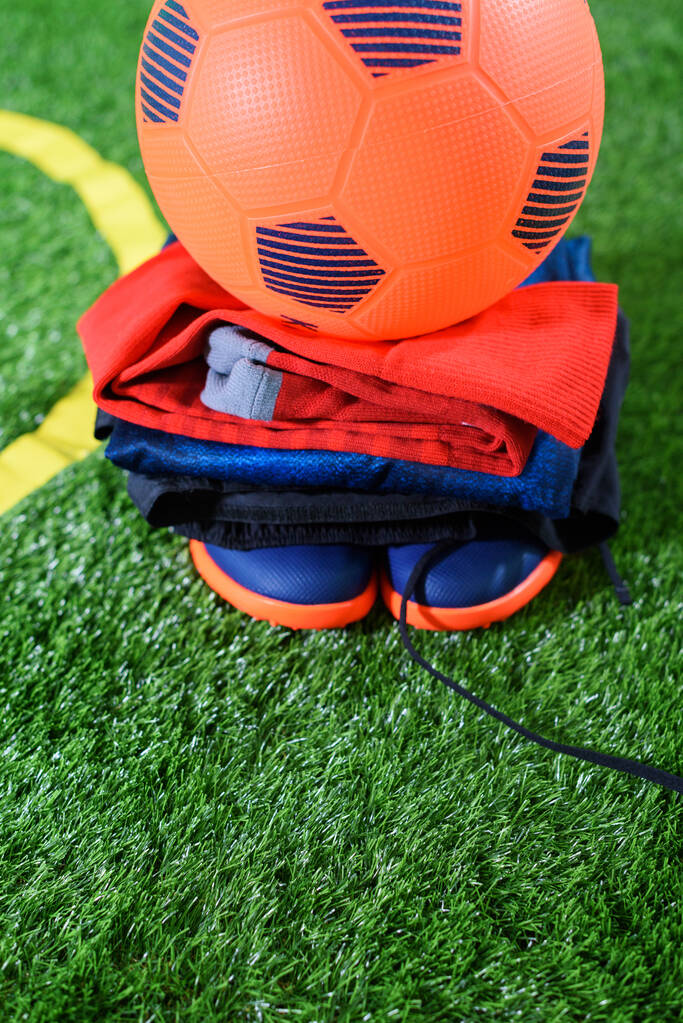 Orange football ball and uniform with shoes on green field kids zone near TV screen at home. Карантин. Сиди дома. Спортивные мероприятия. Коронавирусная ситуация. - Фото, изображение