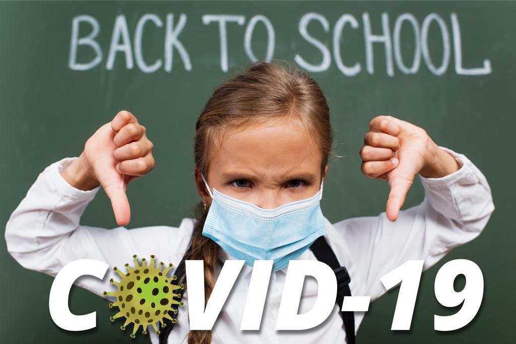 селективный фокус недовольной школьницы в медицинской маске, показывающей большие пальцы у доски с надписью и надписью covid-19 в классе  - Фото, изображение