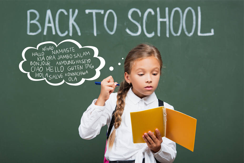 geconcentreerd schoolmeisje met kopieerboek en pen in de buurt van schoolbord en begroeting belettering in de klas  - Foto, afbeelding