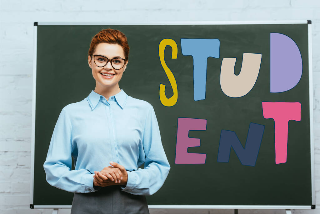 teacher in eyeglasses looking at camera while standing with clenched hands near chalkboard with student lettering  - Photo, Image
