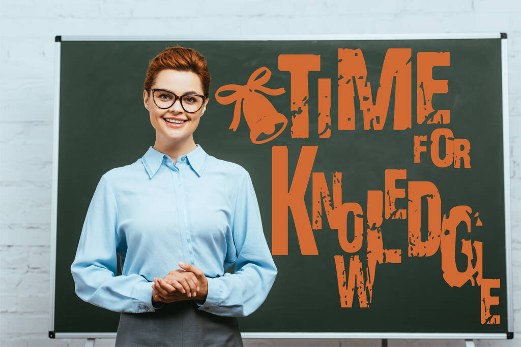 teacher in eyeglasses looking at camera while standing with clenched hands near chalkboard with time for knowledge lettering  - Photo, Image