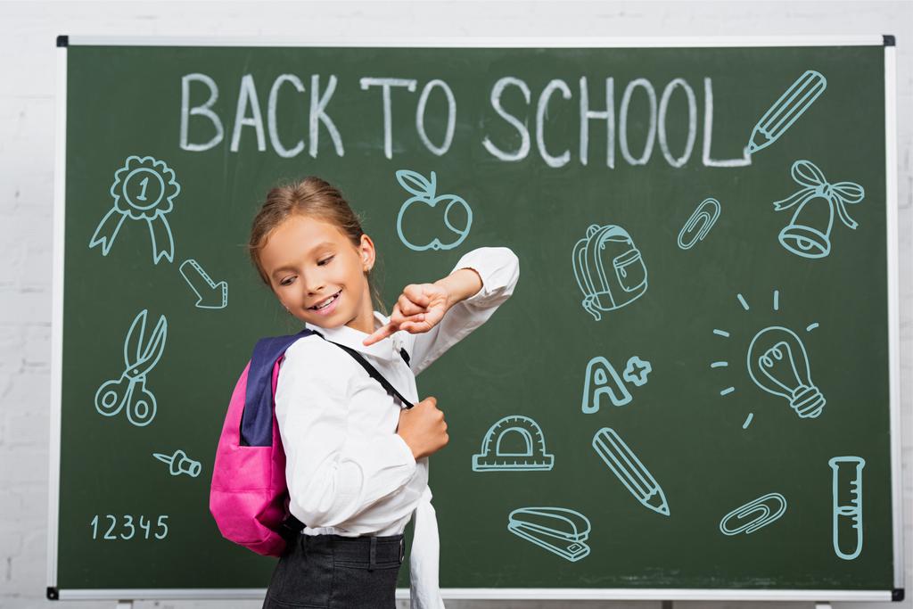 μαθήτρια δείχνοντας με το δάχτυλο στο σακίδιο κοντά στο chalkboard με πίσω στο σχολείο γράμματα και εικονογράφηση  - Φωτογραφία, εικόνα