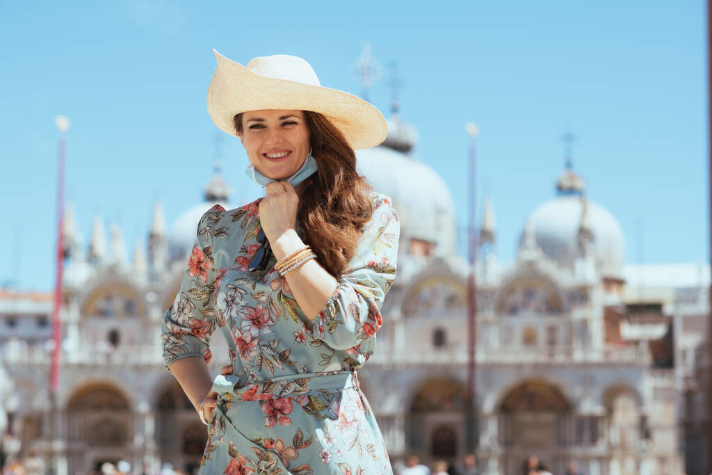 Utazás a covid-19-es járvány idején. boldog elegáns nő virágos ruhában orvosi maszkkal kirándulást a San Marco téren Velence, Olaszország. - Fotó, kép