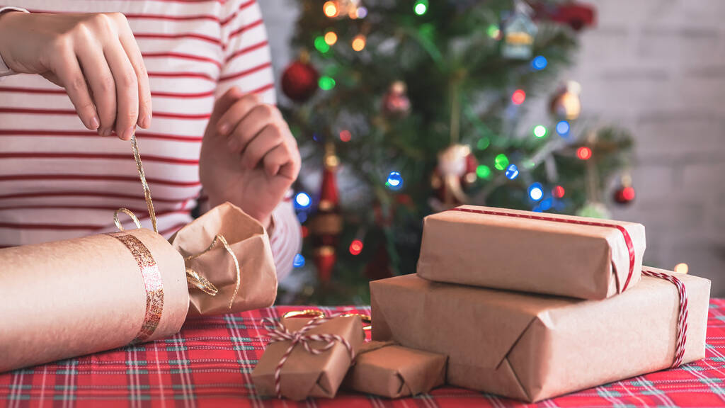 Vrouw verpakking handgemaakte kerstcadeaus voor familie zitten aan tafel met kerstboom met speelgoed en een gloeiende bloemenslinger met bokeh. DIY. - Foto, afbeelding