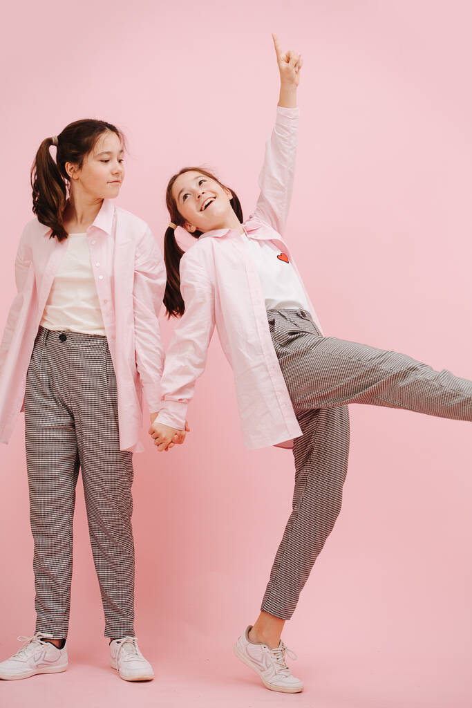 Beautiful little twin sisters happily playing. Over pink background, studio shot. Holding hands. One is artistically walking, looking at the other. Wearing pink dress shirts and checkered pants. - Photo, Image