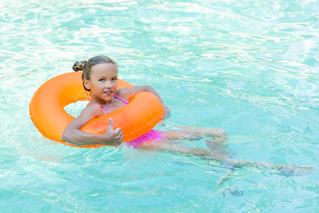 potěšená dívka plovoucí v bazénu na nafukovací kroužek a ukazující palec nahoru - Fotografie, Obrázek