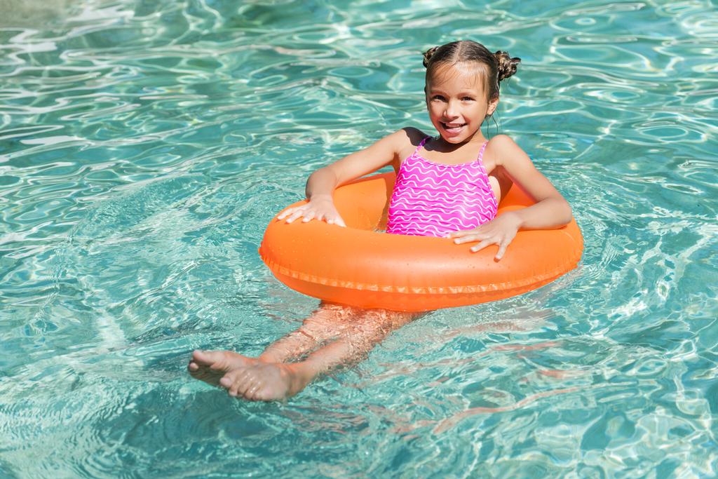 Mädchen blickt in Kamera, während sie auf aufblasbarem Ring im Schwimmbad schwimmt - Foto, Bild