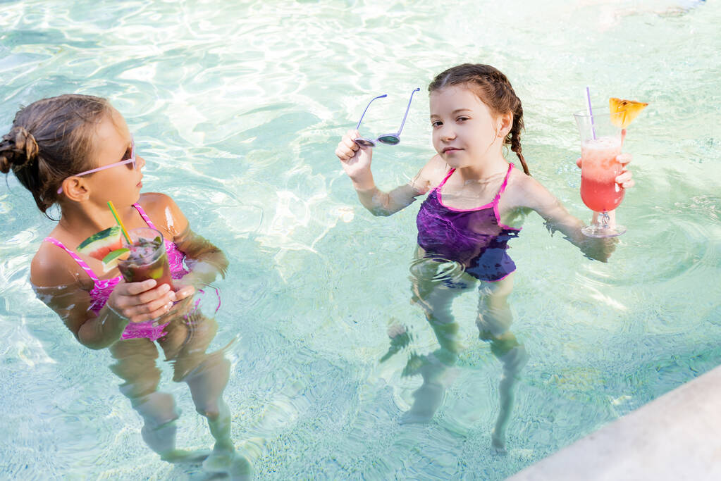 chica mirando a la cámara mientras sostiene gafas de sol y cóctel de frutas en la piscina cerca de amigo - Foto, imagen