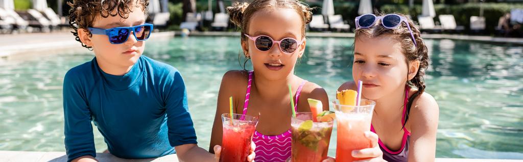 concepto horizontal de niño y niñas en trajes de baño tintineo vasos con refrescantes cócteles cerca de la piscina - Foto, imagen