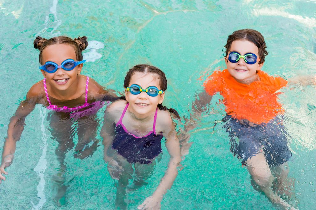 wysoki kąt widzenia dziewcząt i chłopca w goglach pływackich patrząc w kamerę podczas zabawy w basenie - Zdjęcie, obraz