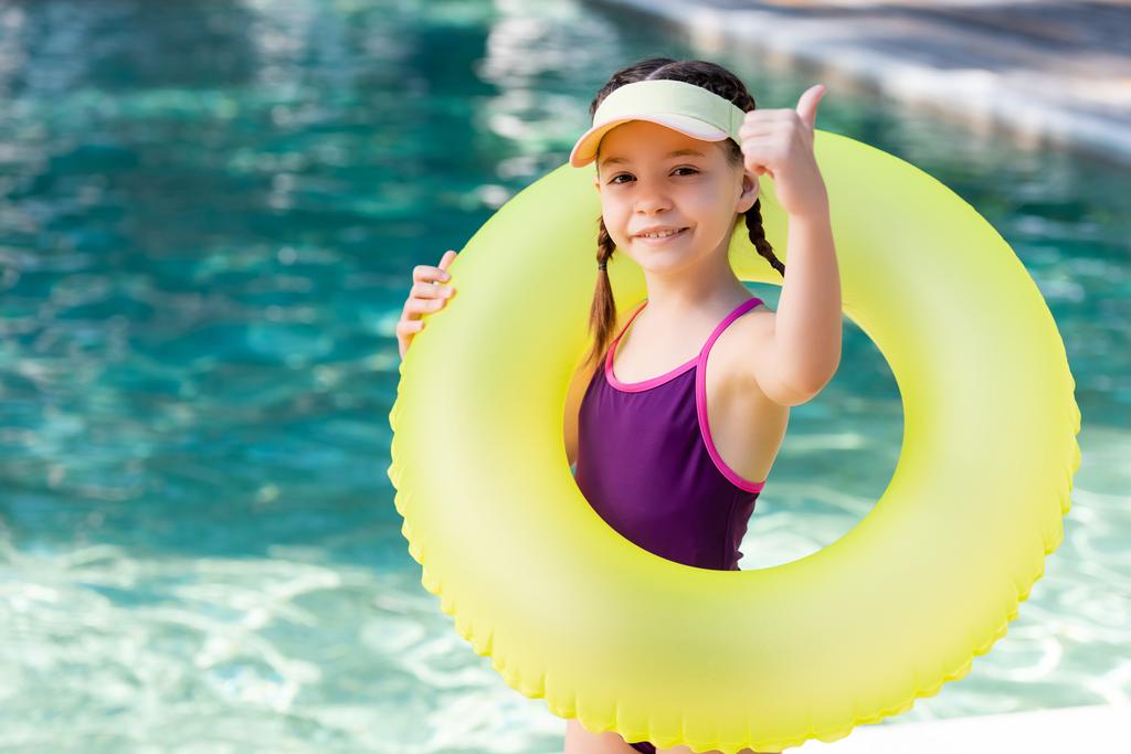 дівчина в купальнику і ковпачок для сонця показує великий палець вгору, тримаючи плавальний кільце біля басейну
 - Фото, зображення