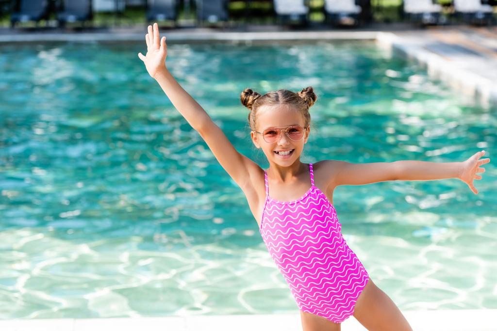 dívka v plavkách a slunečních brýlích stojící s nataženýma rukama u bazénu - Fotografie, Obrázek