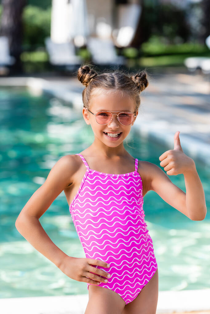 дівчина в купальнику і сонцезахисних окулярах тримає руку на стегні і показує великий палець вгору
 - Фото, зображення