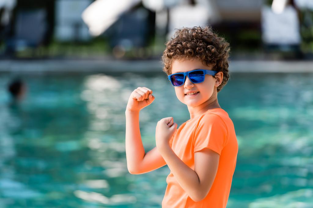 niño rizado en camiseta naranja y gafas de sol demostrando fuerza mientras mira a la cámara - Foto, Imagen