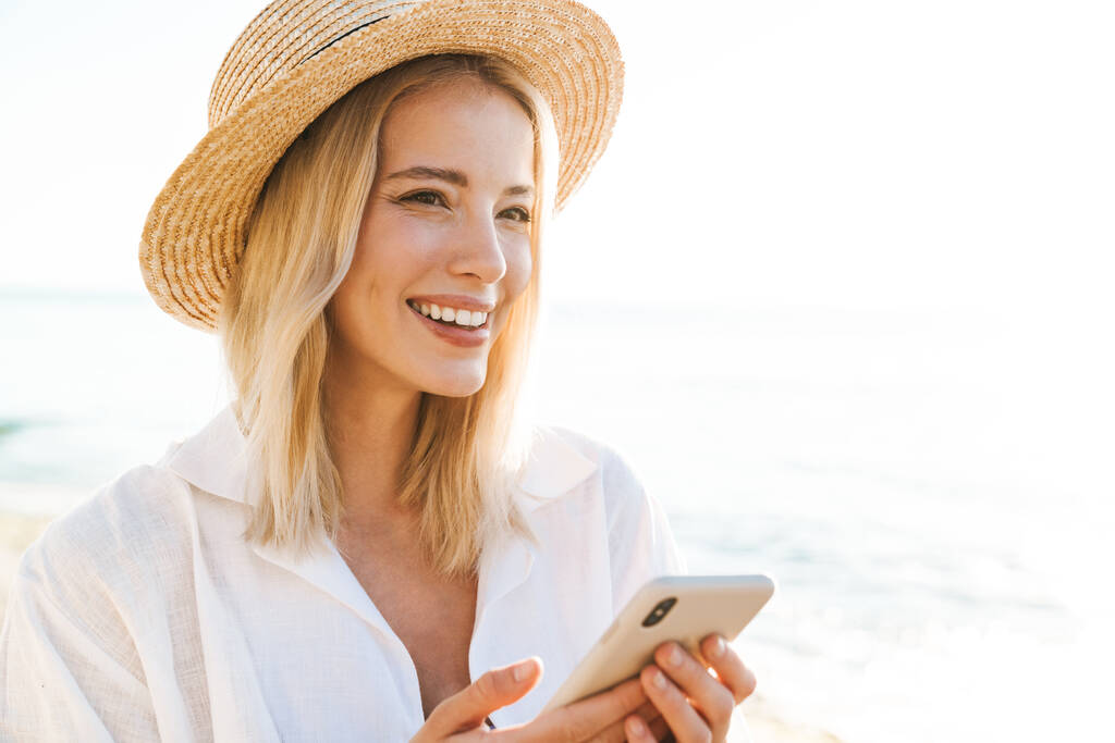 夏のビーチに立っている間、携帯電話を使用して陽気なブロンドの女性のイメージ - 写真・画像
