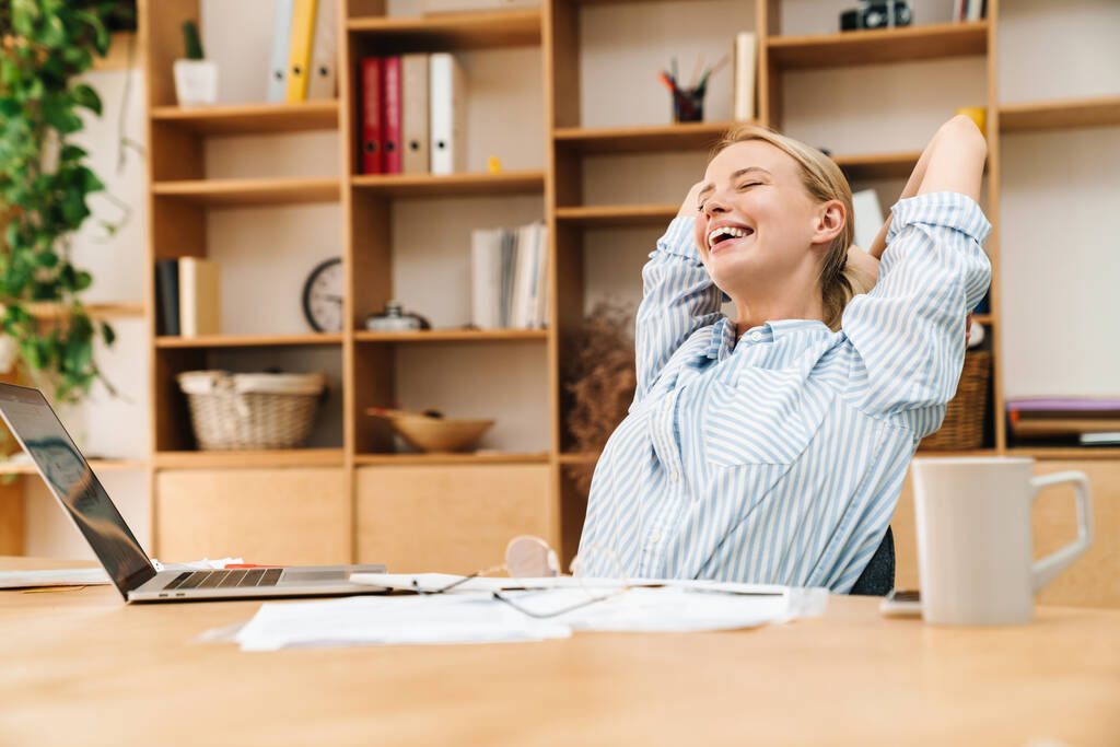 Image of laughing blonde woman stretching her body while working with papers and laptop at table in office - Photo, Image