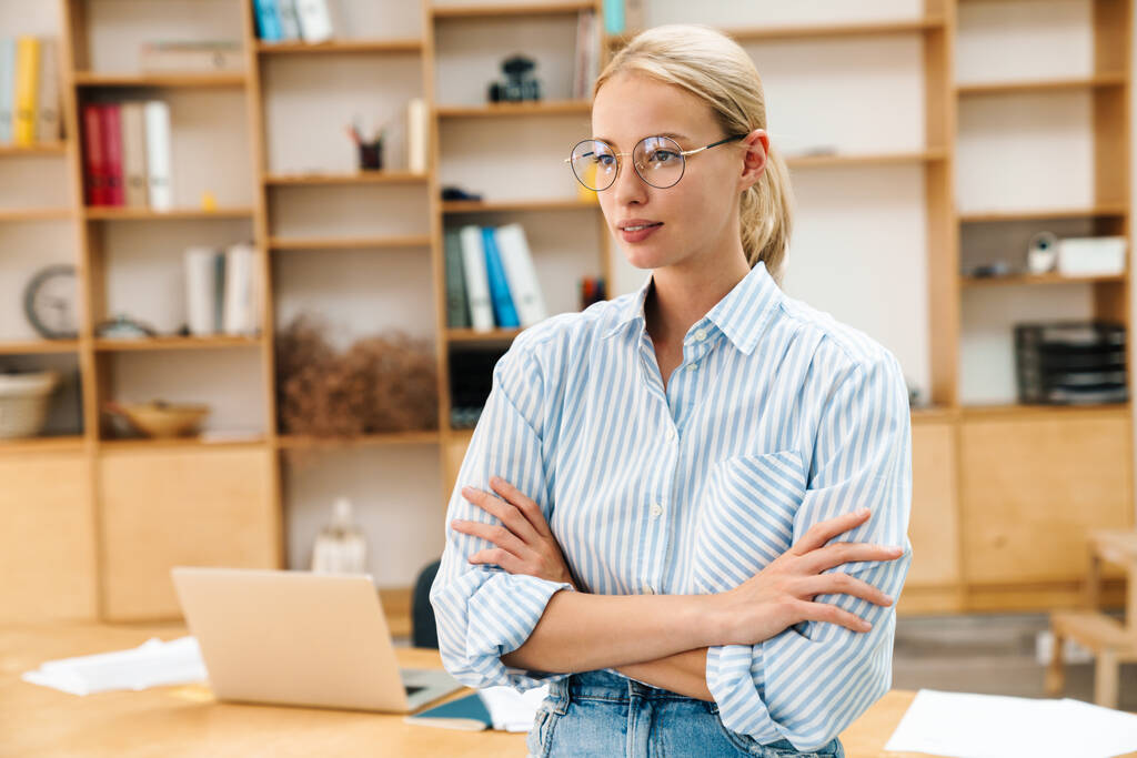 Bild einer jungen attraktiven Frau mit Brille, die mit im Büro verschränkten Händen posiert - Foto, Bild