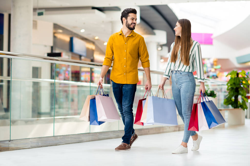 Ganztägiges Foto von positiven fröhlichen Ehepaar Mann Frau fertig einkaufen im Zentrum Mall gehen Spaziergang tragen viele Taschen Pakete tragen gestreifte gelbe Hemd Jeans - Foto, Bild