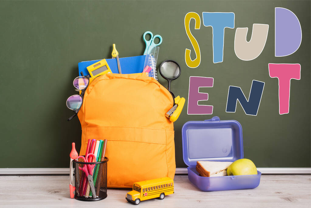 Желтый рюкзак со школьными принадлежностями рядом с коробкой для ланча, игрушечный школьный автобус и держатель ручки на столе рядом с зеленой доской с надписью  - Фото, изображение