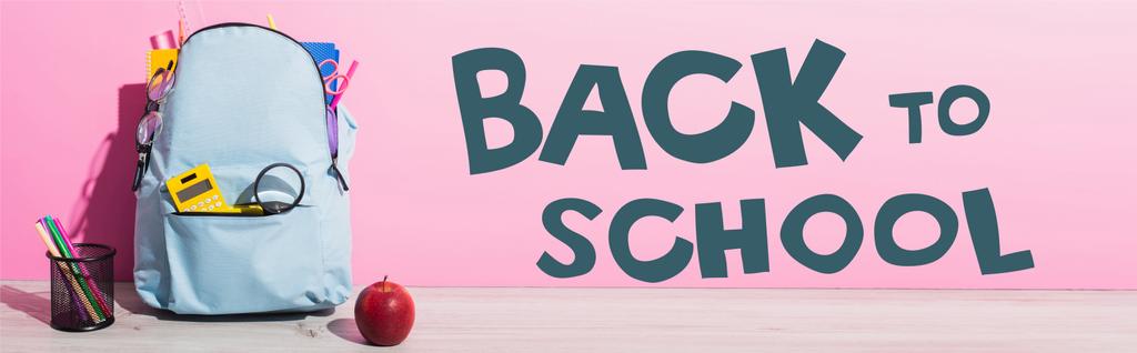 immagine orizzontale di zaino blu con materiale scolastico vicino mela intera, portapenne con pennarelli e iscrizione a scuola su rosa - Foto, immagini