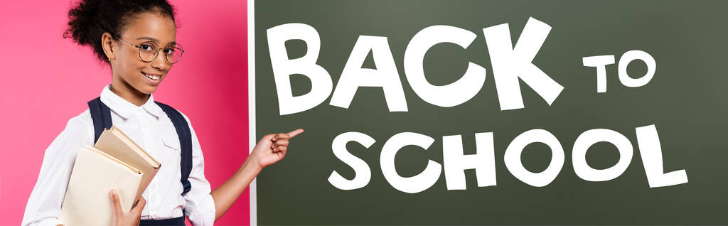 african american schoolgirl pointing with finger at chalkboard with back to school lettering on pink, panoramic shot - Photo, Image