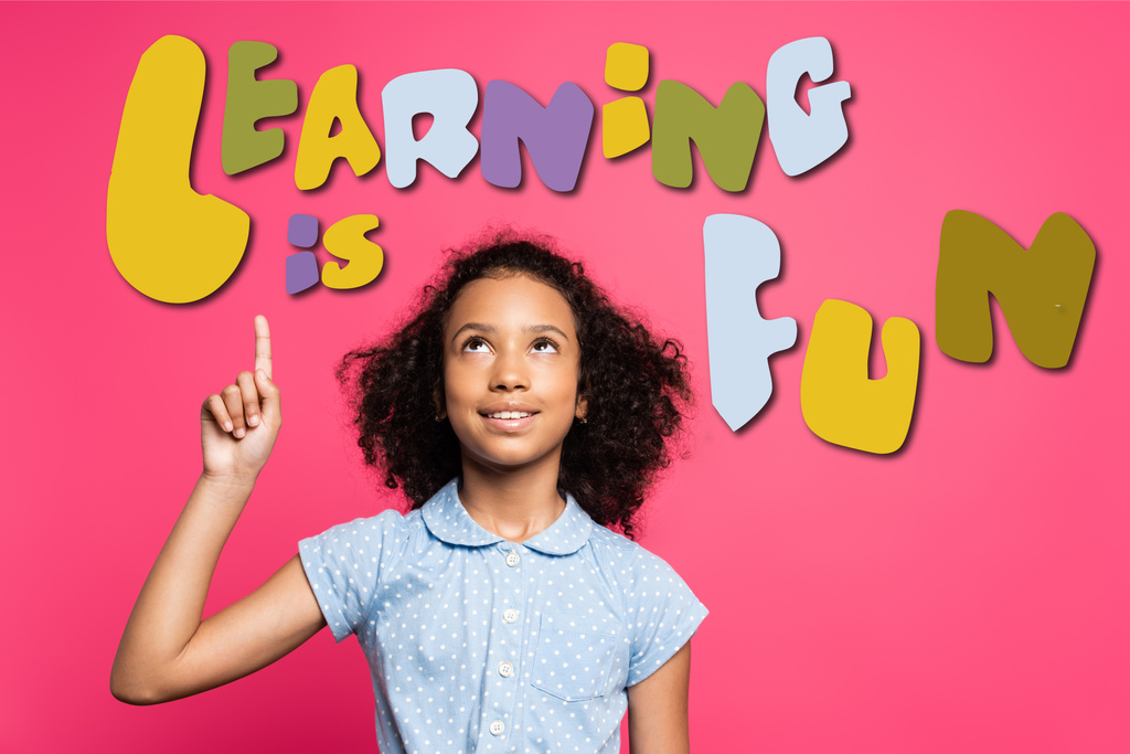 rizado africano americano niño apuntando con el dedo y mirando hacia arriba cerca de aprender es divertido letras en rosa - Foto, imagen