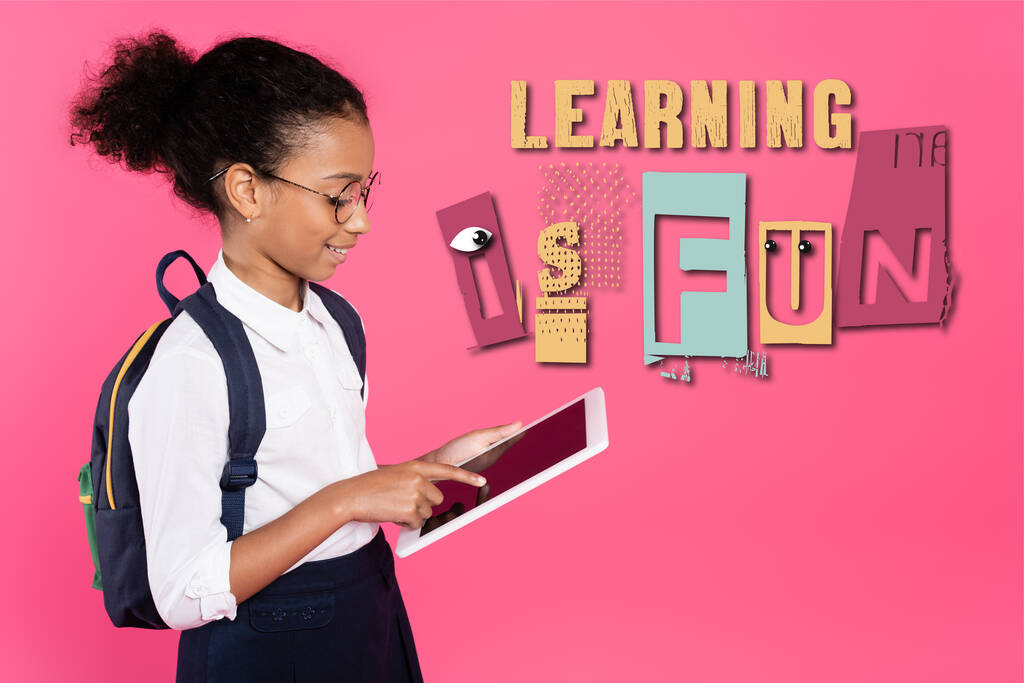 Afričanky americká školačka v brýlích s batohem pomocí digitálního tabletu v blízkosti učení je zábavné písmo na růžové - Fotografie, Obrázek