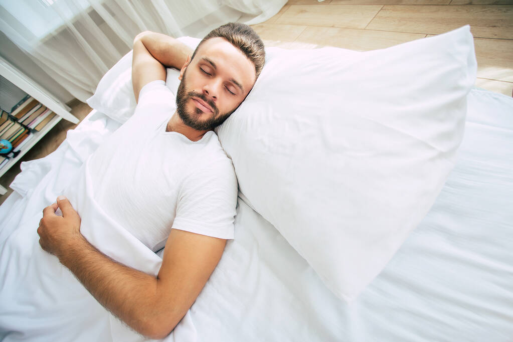 Beau jeune homme barbu dort dans un grand lit blanc dans une chambre confortable - Photo, image