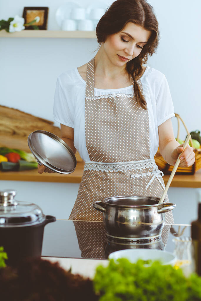 Giovane donna bruna che cucina zuppa in cucina. Casalinga che tiene in mano un cucchiaio di legno. Concetto alimentare e sanitario - Foto, immagini