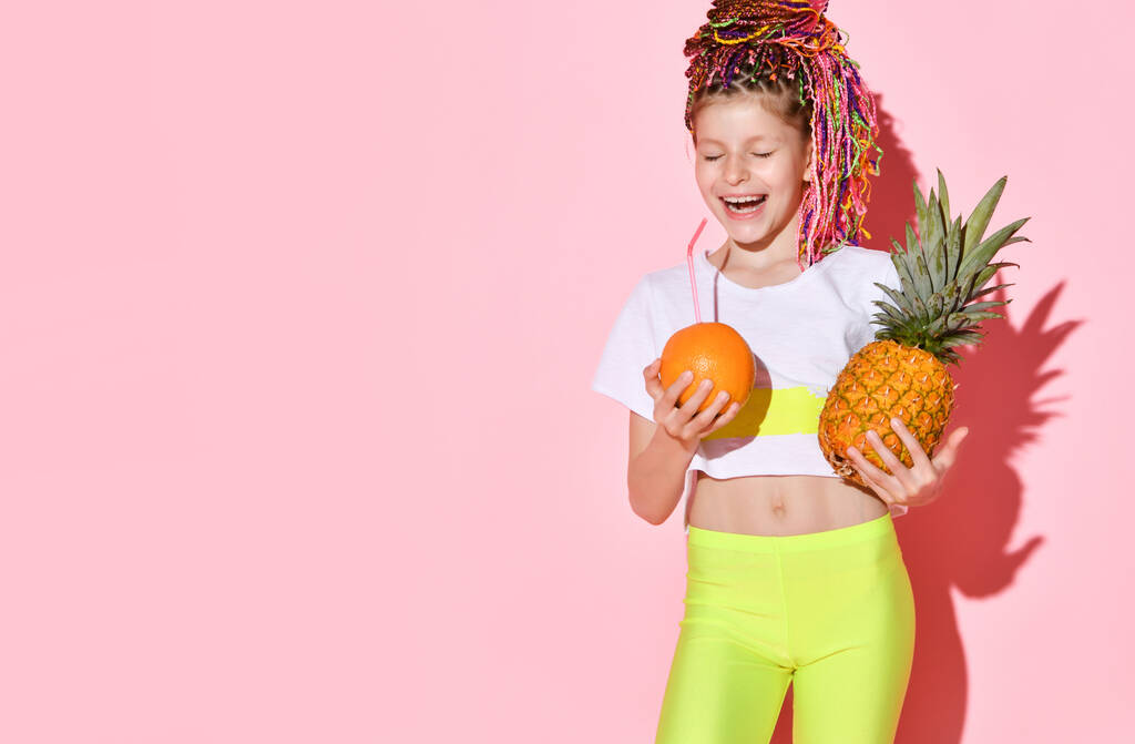 Niza niña con cornalinas multicolores en la cabeza riendo alegremente con los ojos cerrados sosteniendo piña y naranja en las manos - Foto, Imagen