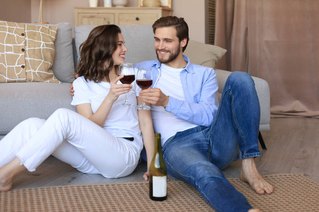 Happy couple sitting, relaxing on floor in living room, drinking red wine. Smiling young husband and wife rest at home enjoy romantic date on family weekend together. - Photo, Image