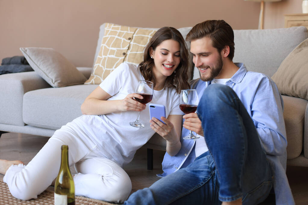 Szczęśliwa para siedzi, relaksuje się na podłodze w salonie, pije czerwone wino, patrzy razem na smartfona. Uśmiechnięty młody mąż i żona odpoczynku w domu cieszyć się romantyczną randkę na rodzinny weekend razem - Zdjęcie, obraz