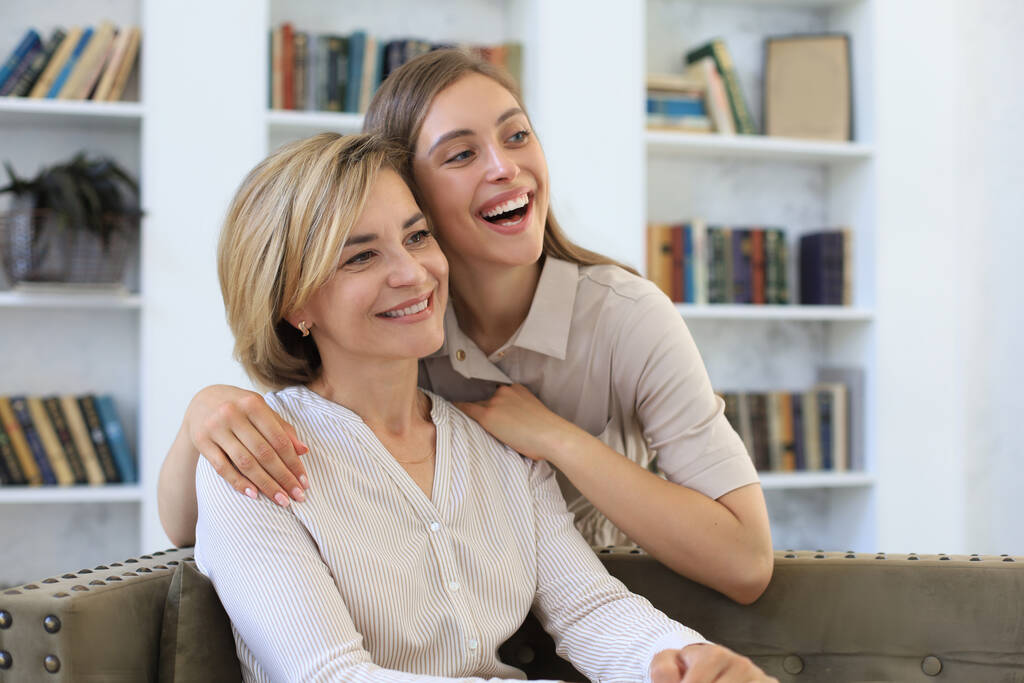 Красивая мама средних лет и ее взрослая дочь обнимаются и улыбаются, сидя дома на диване - Фото, изображение