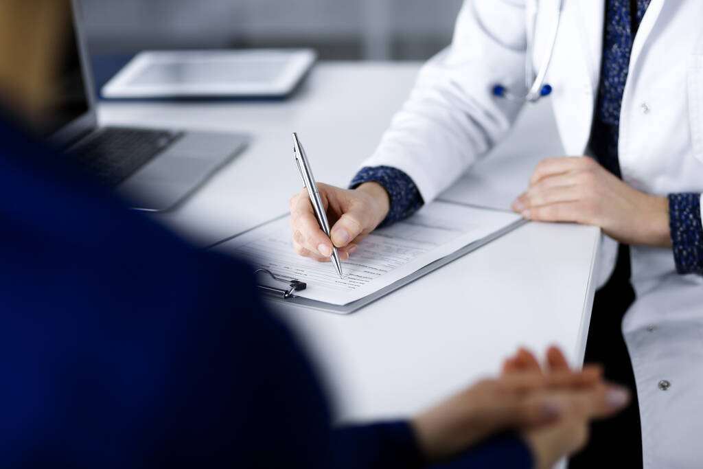 Невідома жінка-лікар пише деякі медичні рекомендації для свого пацієнта, поки вони сидять разом за столом у кабінеті в клініці. Лікар використовує кишеню, крупним планом. Ідеальний
 - Фото, зображення