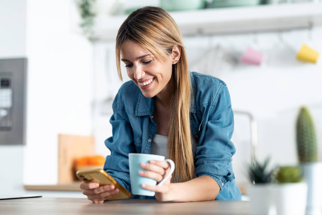 Tiro de mujer joven sonriente usando su teléfono móvil mientras bebe una taza de té en la cocina en casa. - Foto, Imagen
