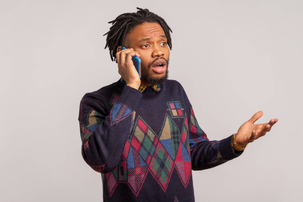 Обеспокоенный бородатый африканский парень с дредами, говорящий по телефону с открытым ртом и большими глазами, шокированный разочарованной информацией. Крытая студия, снятая на сером фоне - Фото, изображение