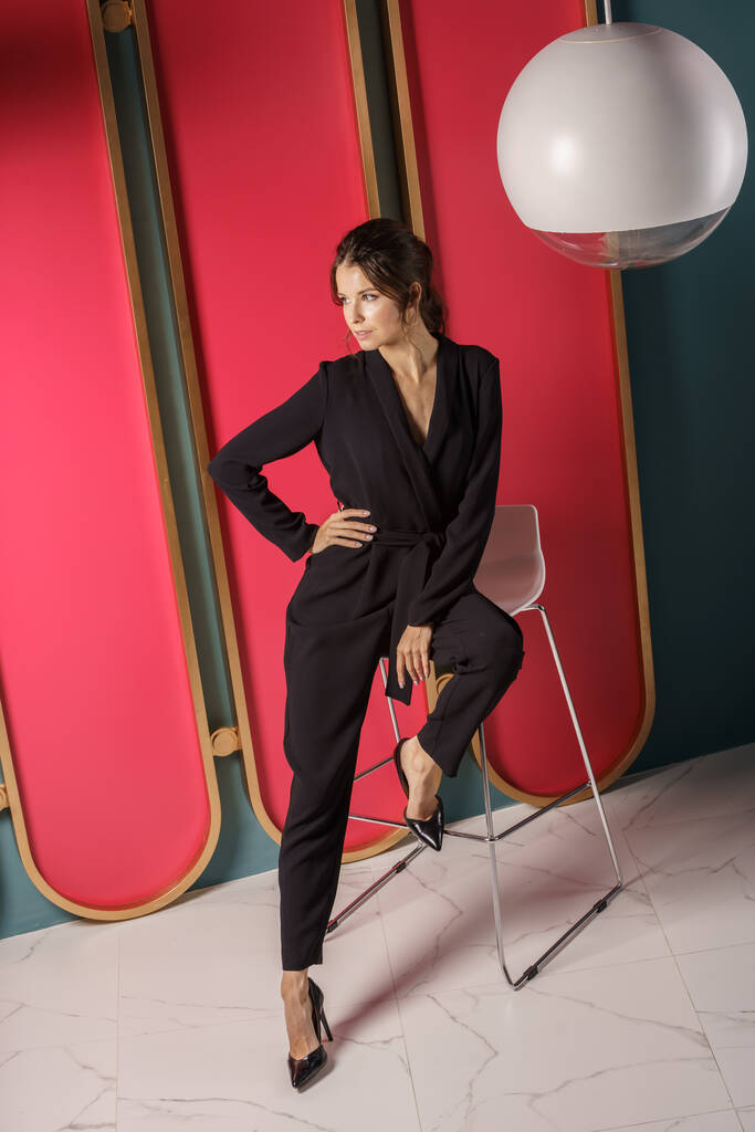 Предпринимательница стоит у красной стены в интерьере роскошных квартир. Портрет брюнетки в черном костюме и на высоких каблуках  - Фото, изображение
