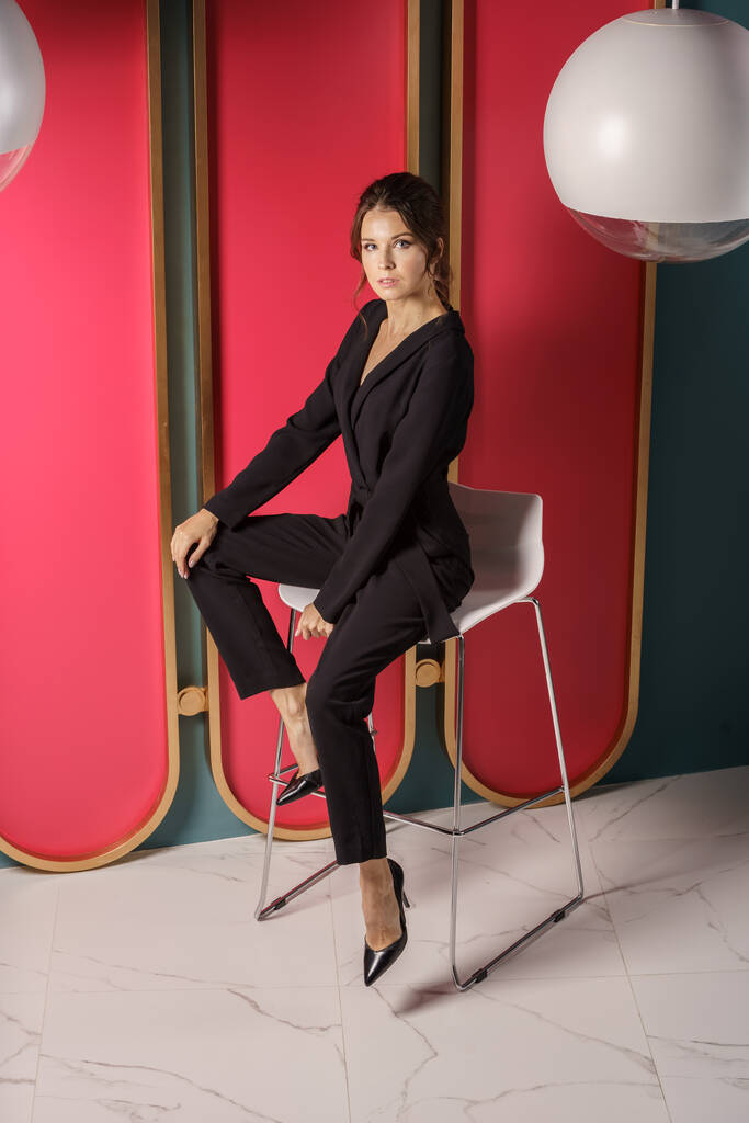 Предпринимательница позирует рядом с красной стеной в интерьере роскошных квартир. Портрет брюнетки в черном костюме и на высоких каблуках  - Фото, изображение