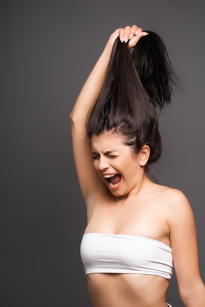 sconvolto bruna donna tenendo i capelli danneggiati e urlando isolato su nero - Foto, immagini