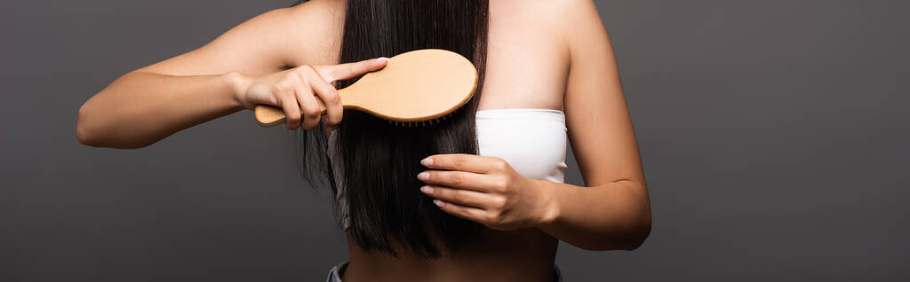 abgeschnittene Ansicht einer brünetten Frau, die glänzendes Haar isoliert auf schwarzem, panoramischem Foto bürstet - Foto, Bild