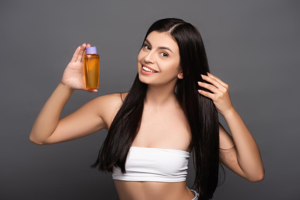 donna bruna che tiene olio per capelli in bottiglia isolato su nero - Foto, immagini