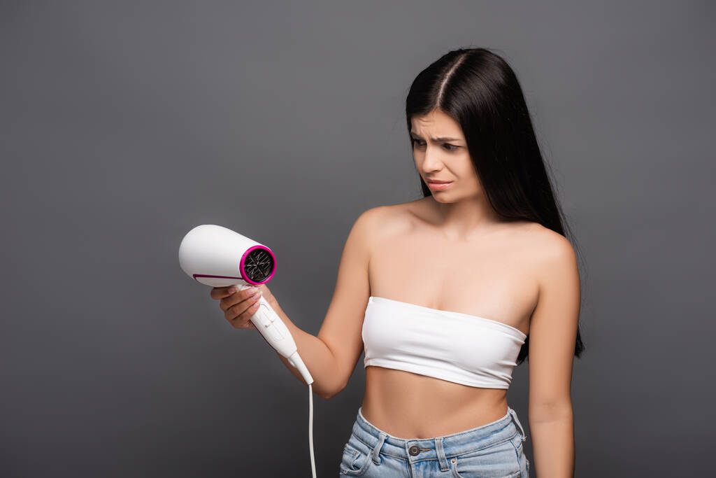 worried brunette woman looking at hair dryer isolated on black - Photo, Image