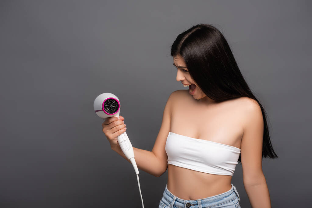 brunette woman looking at hair dryer and yelling isolated on black - Photo, Image