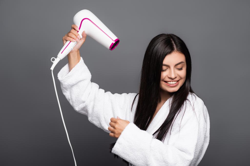 morena de cabelos longos mulher em roupão de banho usando secador de cabelo isolado no preto - Foto, Imagem