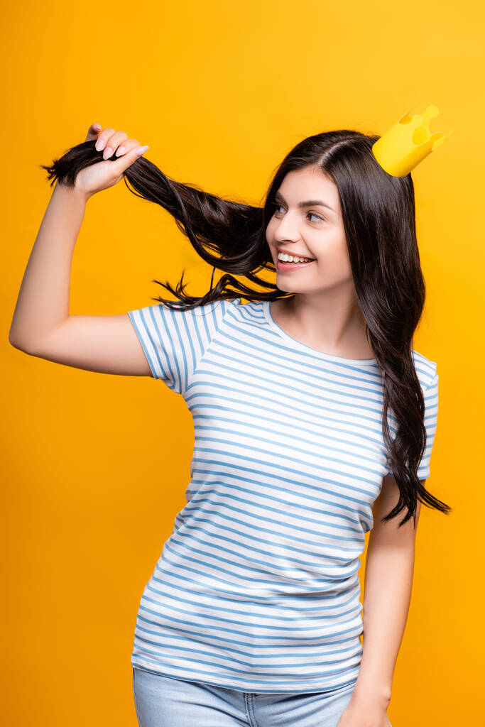 μελαχρινή γυναίκα σε χαρτί στέμμα κρατώντας τα μαλλιά και χαμογελώντας απομονώνονται σε κίτρινο - Φωτογραφία, εικόνα