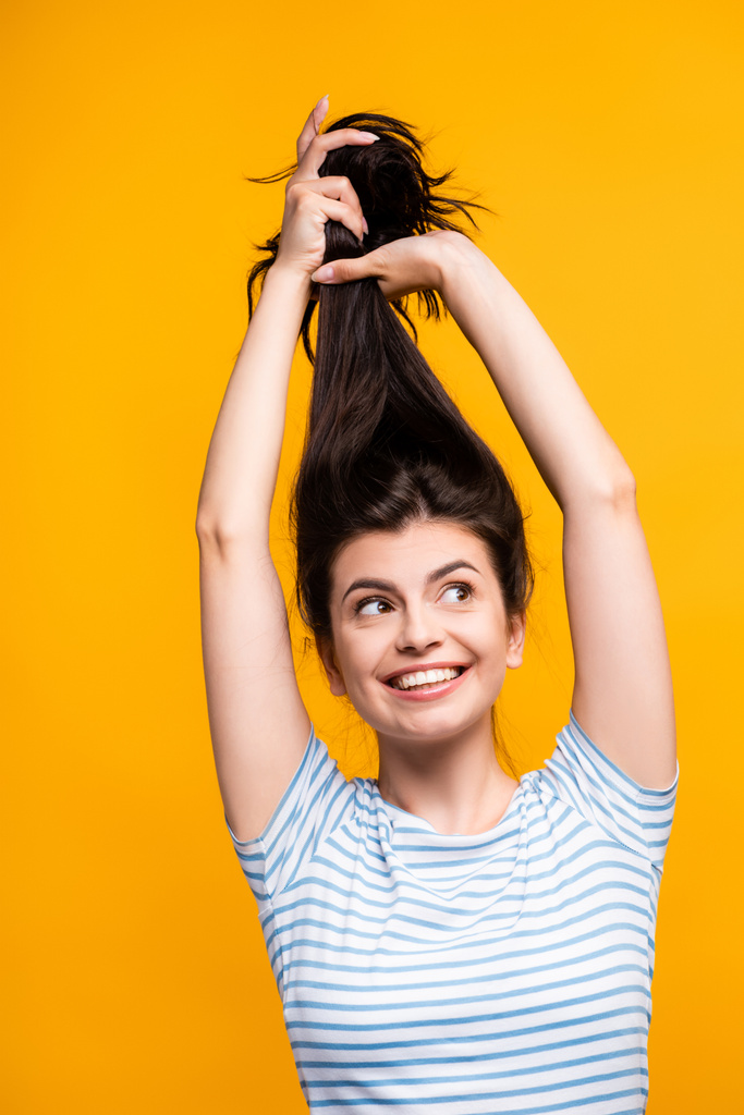 брюнетка женщина держа волосы над головой и улыбаясь изолированы на желтый - Фото, изображение