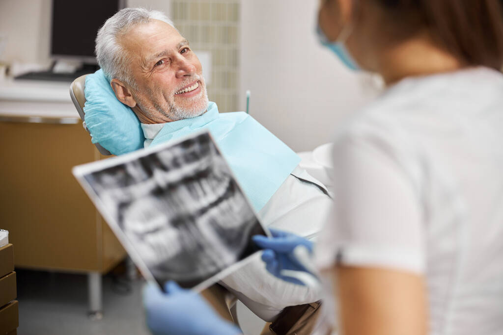 Idős polgár, aki fogorvosi székben ül, és egy fogtechnikusra néz. - Fotó, kép