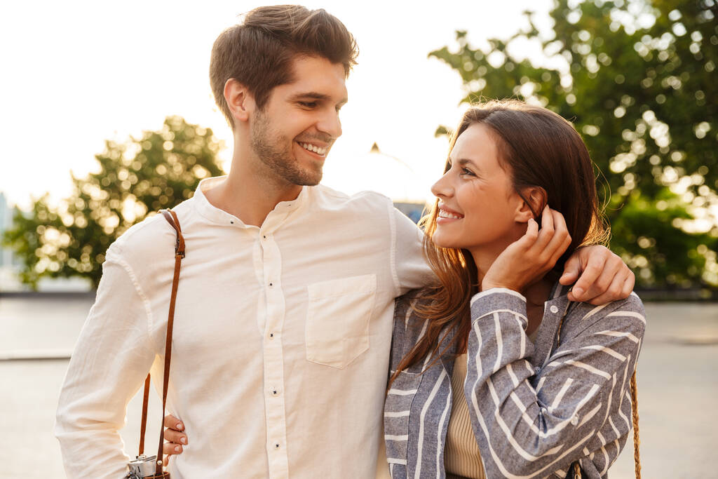 街中をレトロなカメラで歩きながら笑顔で抱きしめる若い白人夫婦の姿 - 写真・画像