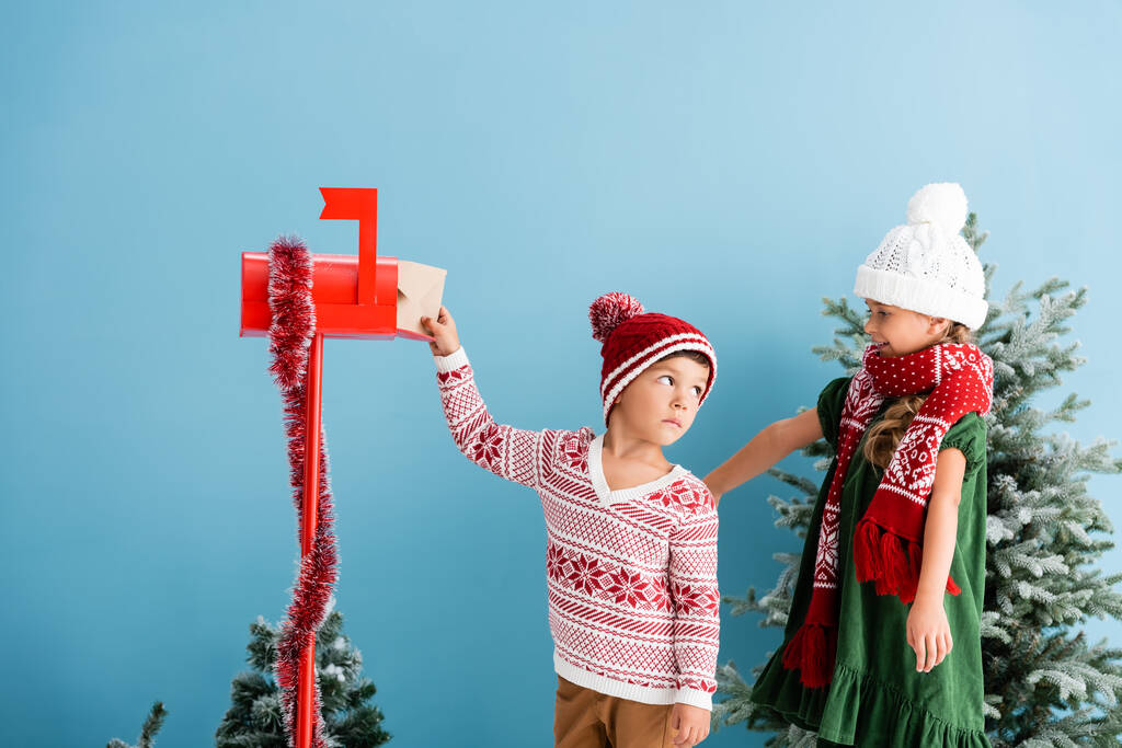 Junge mit Hut und Pullover steckt Umschlag in Briefkasten bei Schwester im Winteroutfit - Foto, Bild