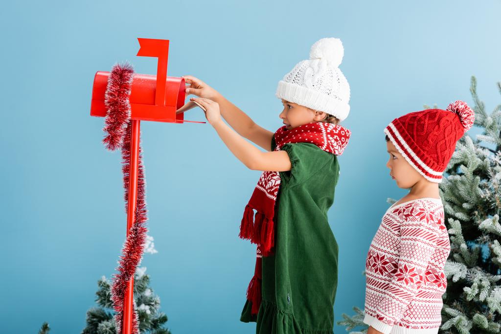 enfant en tenue d'hiver mettre enveloppe dans la boîte aux lettres près frère isolé sur bleu - Photo, image